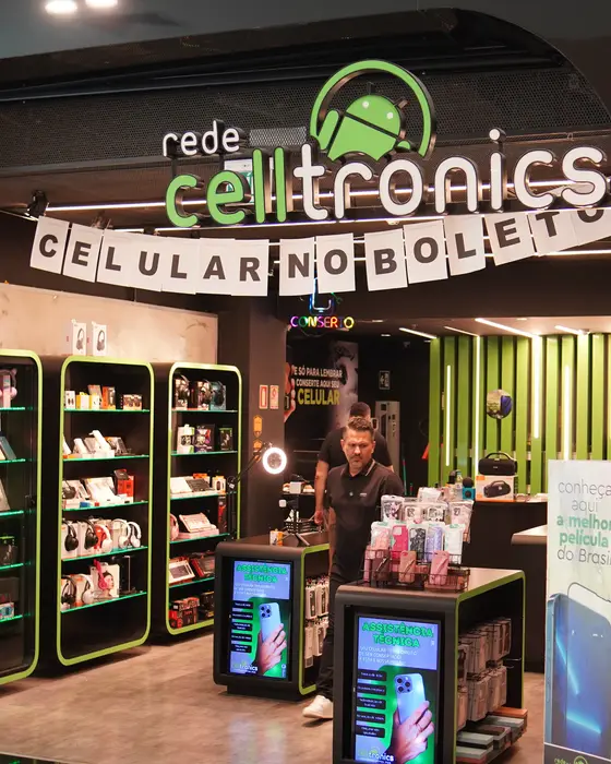 Fachada de loja da Rede Celltronics com parede de acessórios, faixa de celular no boleto e totens de assistência técnica